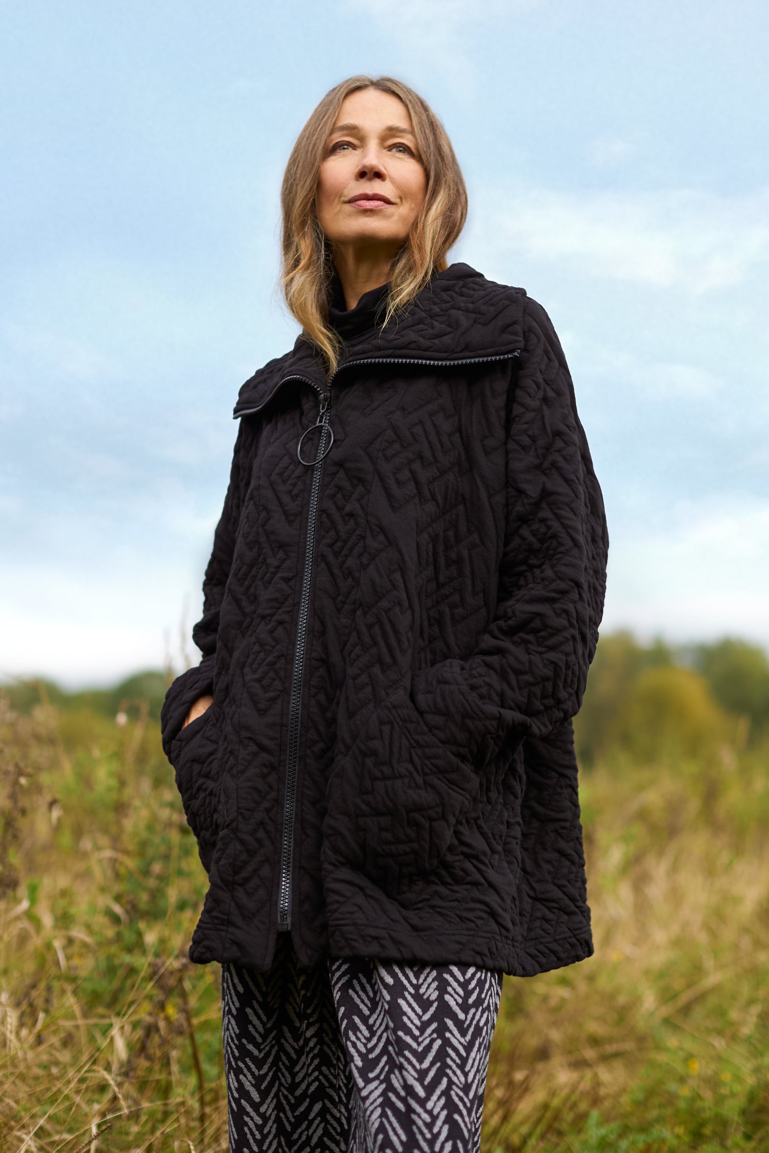 A woman stands outdoors wearing the chic Abstract Quilted Cotton Jacket, featuring dolman sleeves, paired with patterned pants. In the background, a grassy field extends under a cloudy sky, forming a peaceful scene.