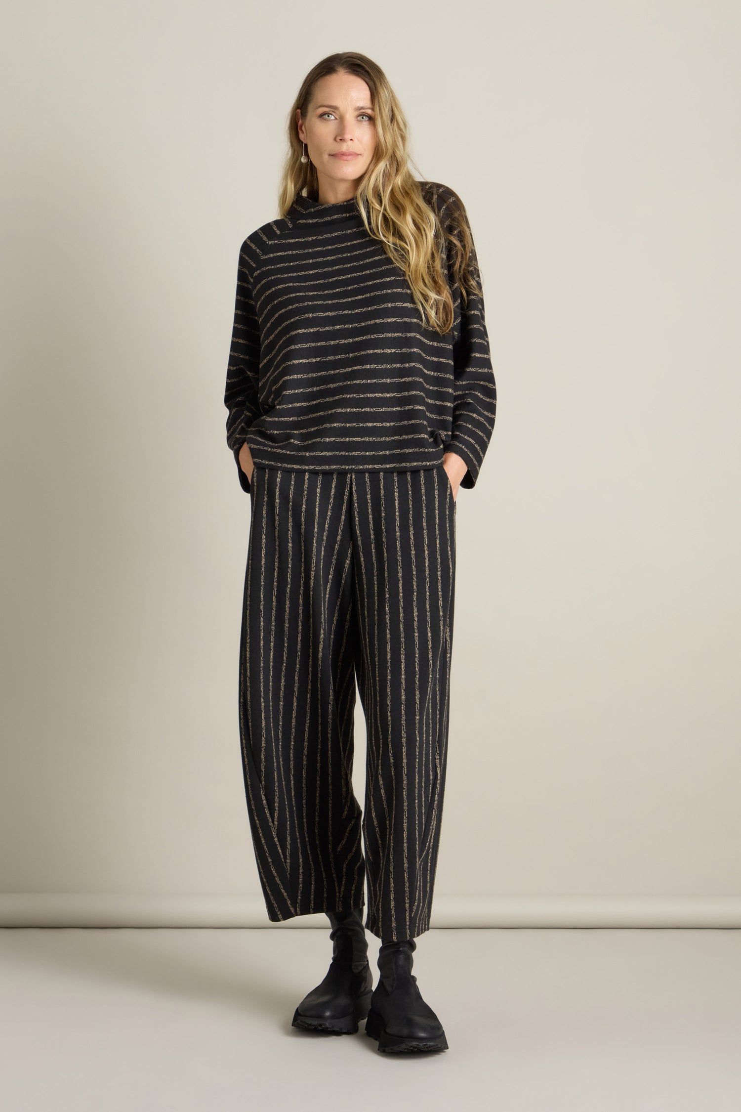 A person stands against a plain background wearing a black, striped top paired with Broken Stripe Jersey Bubble Trousers, both featuring vertical lines. With one hand in their pocket and black shoes completing their look, they exude a modern edge.