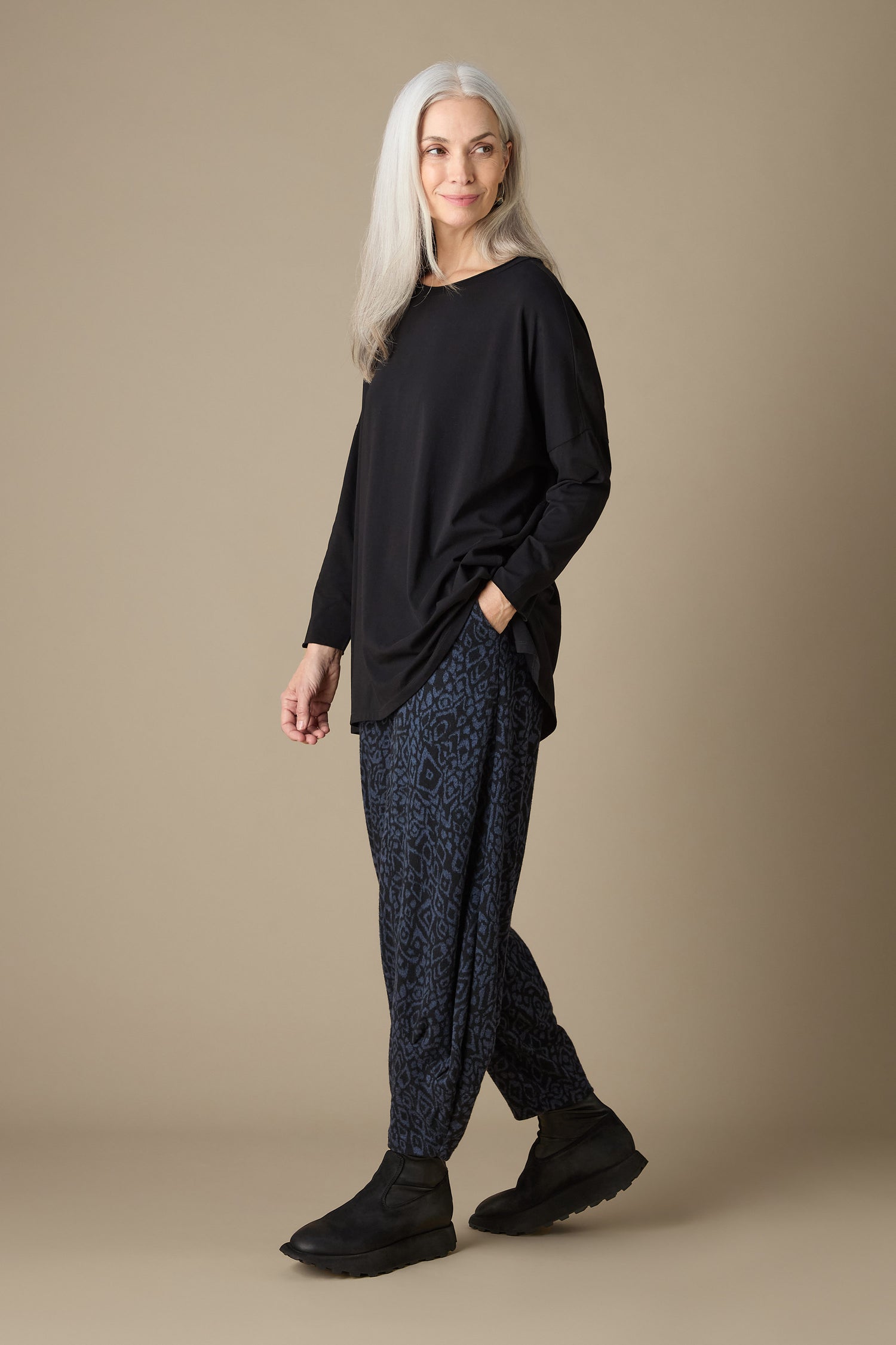 A person with long gray hair is wearing a black long-sleeve top, Ikat Soft Jersey Bubble Trousers in blue and black abstract print cropped length, and black shoes, standing against a beige background.