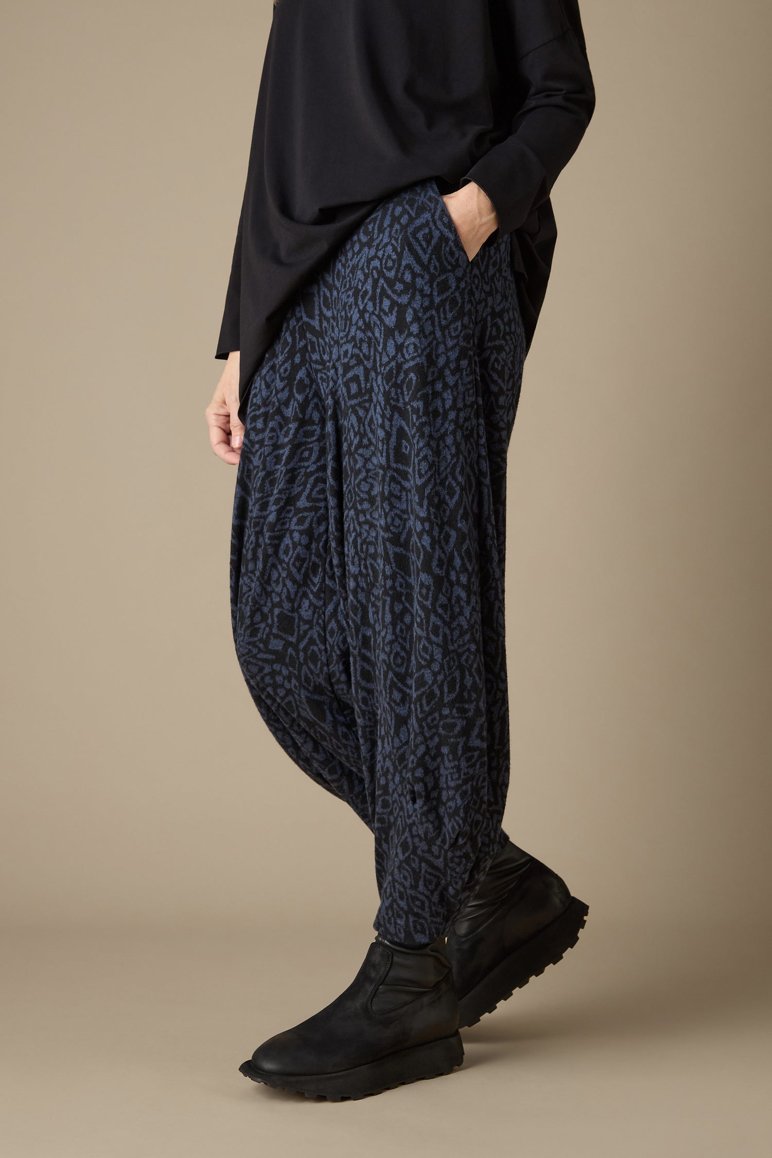 A person wearing a black long-sleeve top and the Ikat Soft Jersey Bubble Trouser featuring a blue and black abstract print stands against a neutral background, accentuated by their black shoes.