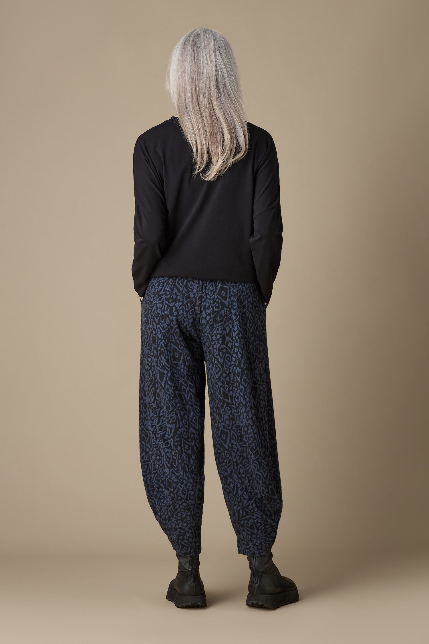 A person with long gray hair, wearing a black sweater and the Ikat Soft Jersey Bubble Trouser featuring a blue and black abstract print, stands against a plain background in cropped-length trousers, facing away from the camera.