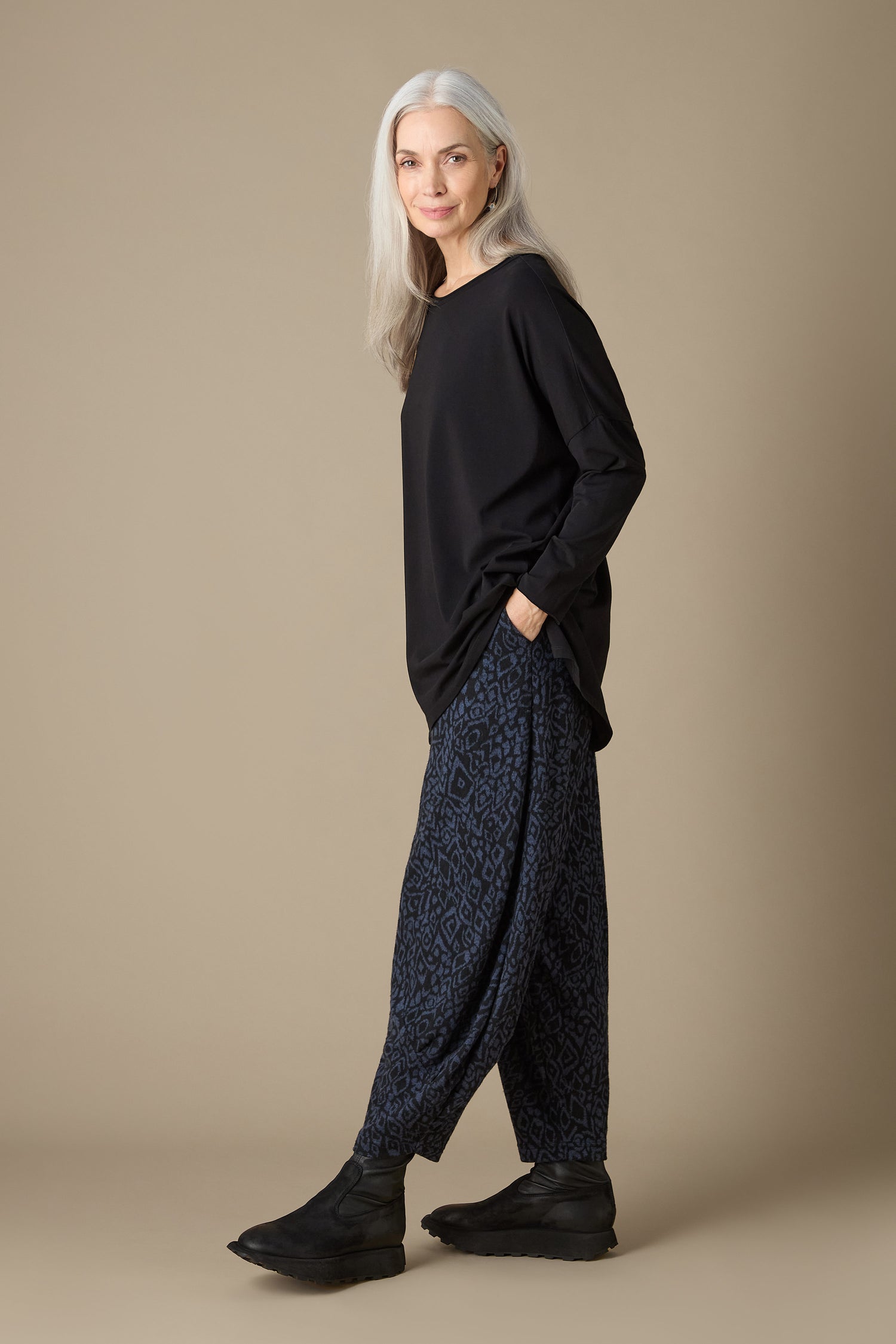 A person with long gray hair wears a black top and cropped Ikat Soft Jersey Bubble Trousers adorned with a blue and black abstract print, paired with black shoes, as they stand against a neutral background.