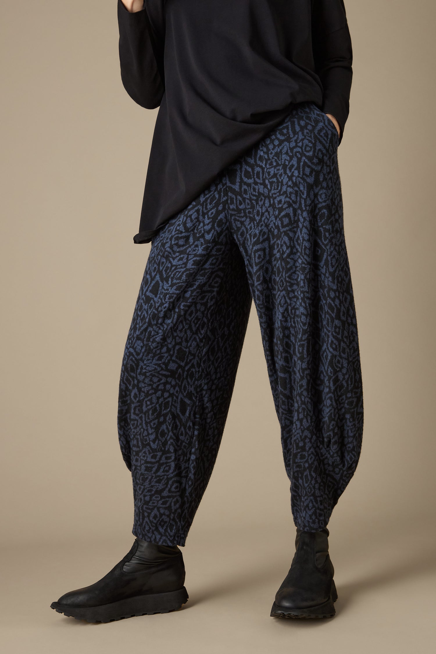 The individual is dressed in Ikat Soft Jersey Bubble Trousers featuring a blue and black abstract print, paired with a black top and black boots, set against a neutral backdrop.