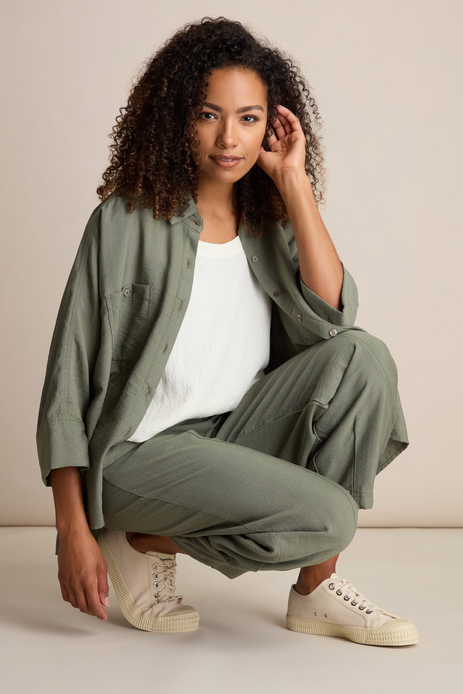 A woman with curly hair, dressed in a green button-up shirt, white top, and transitional Textured Viscose Bubble Trouser with an elasticated waistband. She squats with her left arm resting on her knee and right hand touching her hair. She is wearing beige sneakers.