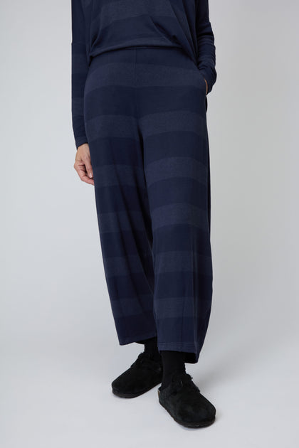 A person wearing Soft Viscose Jersey Stripe Bubble Trouser in dark blue wide stripes, paired with a matching long-sleeve top, black socks and slippers, stands against a plain light background.