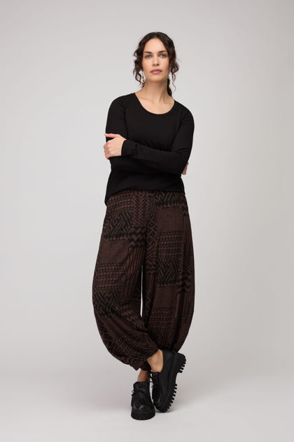 Woman with arms crossed and neutral expression stands against a plain background wearing a black long-sleeve top, black shoes, and Textured Ikat Patchwork Jersey Bubble Trouser featuring an elasticated waist.