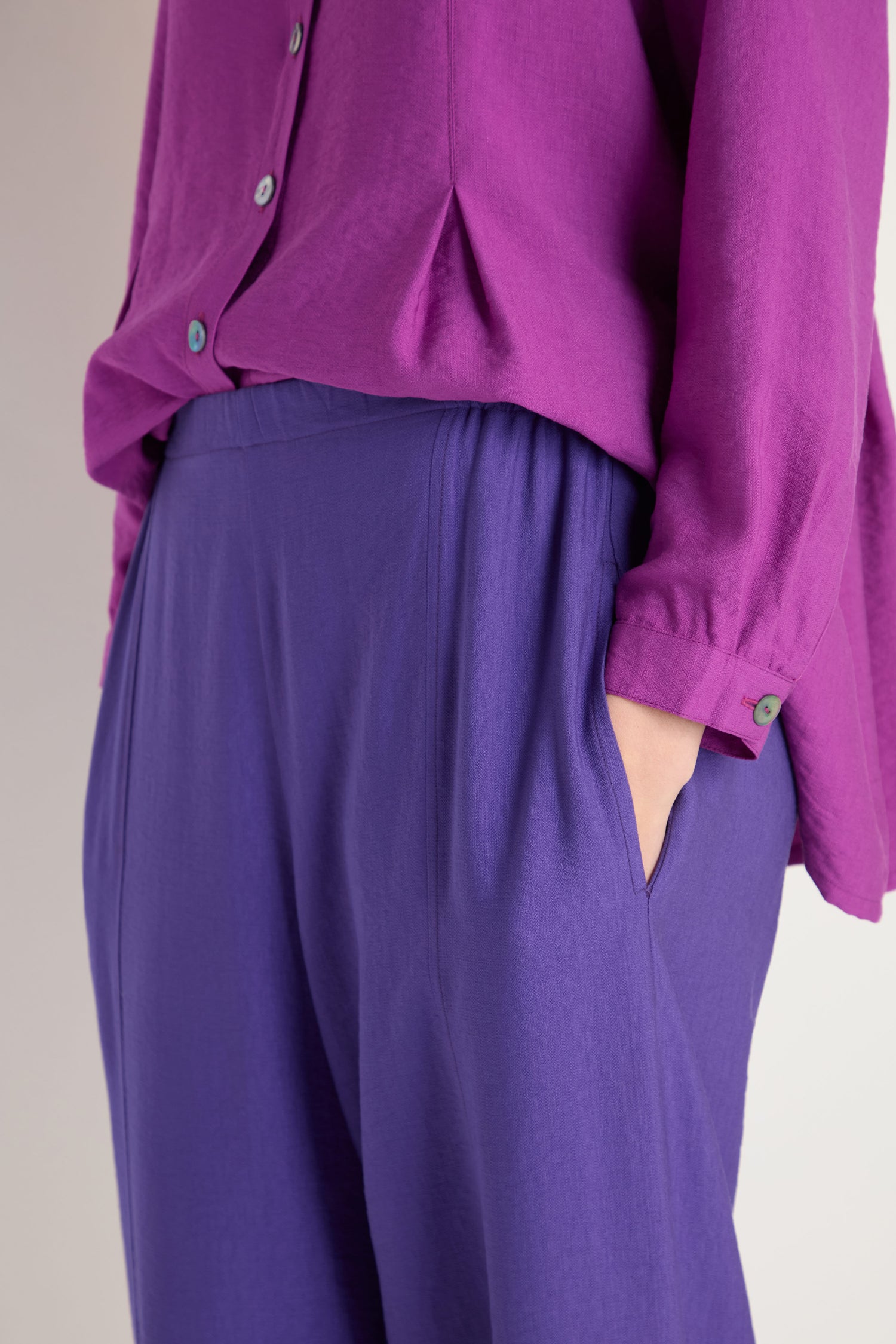 Person wearing Textured Viscose Bubble Trouser—loose-fitting, cropped purple pants—with one hand in the pocket, photographed from the waist down and paired with a long-sleeved magenta shirt.