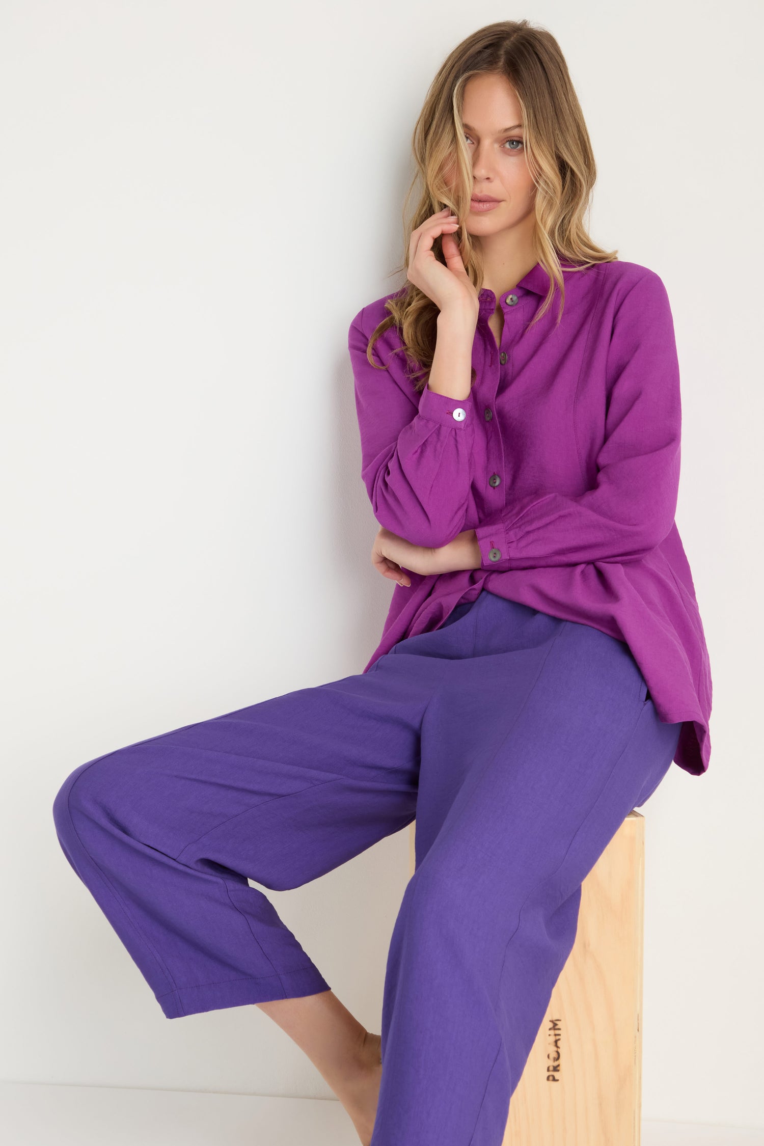 A woman sits on a wooden block wearing the Textured Viscose Bubble Trouser and a loose-fitting purple shirt, with one hand touching her face.