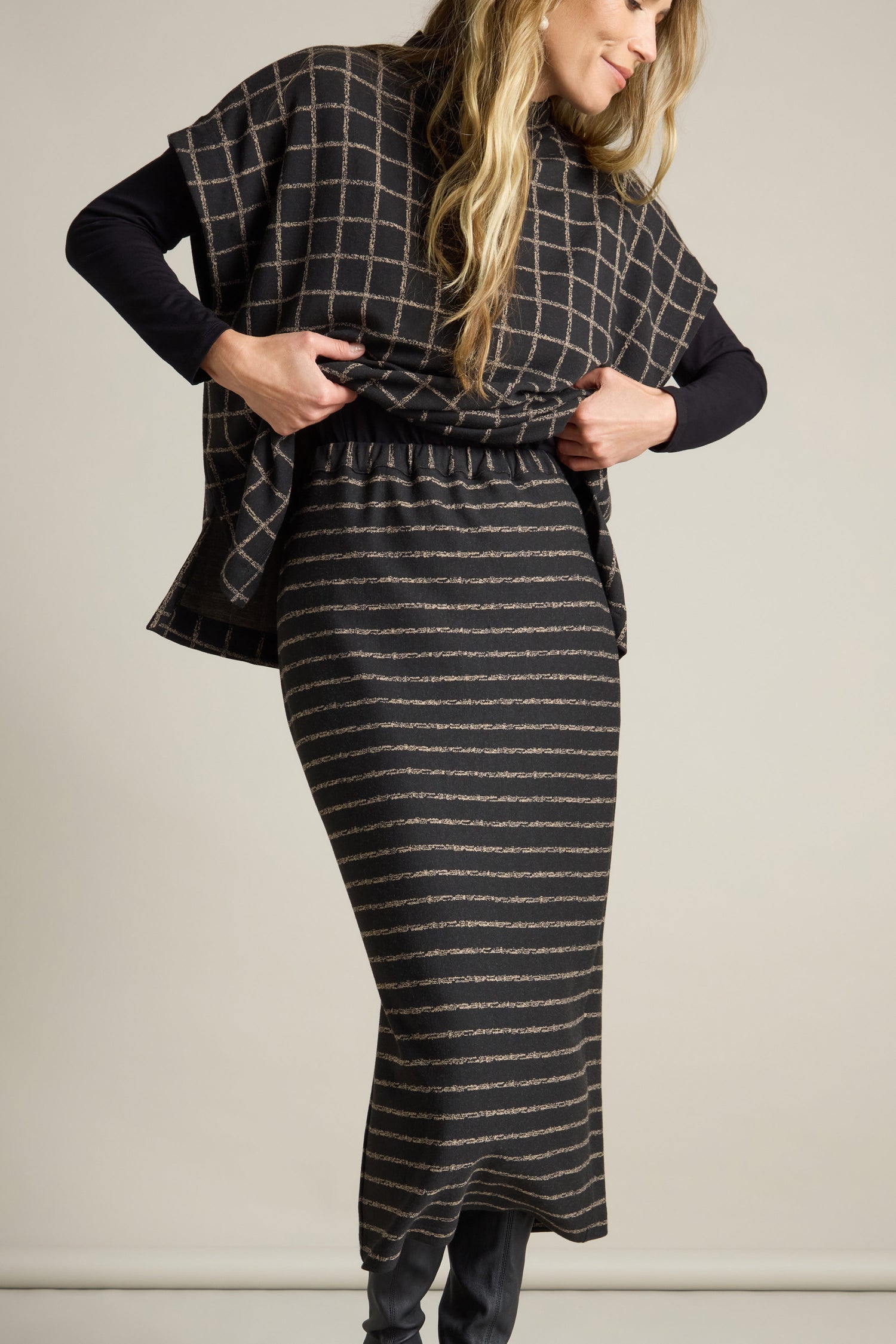 In front of a simple backdrop, an individual in a dark ensemble showcases the Broken Stripe Jersey Tube Skirt, crafted from soft jersey fabric with horizontal stripes, and pairs it with a top featuring vertical checks, adjusting the top with their hands.