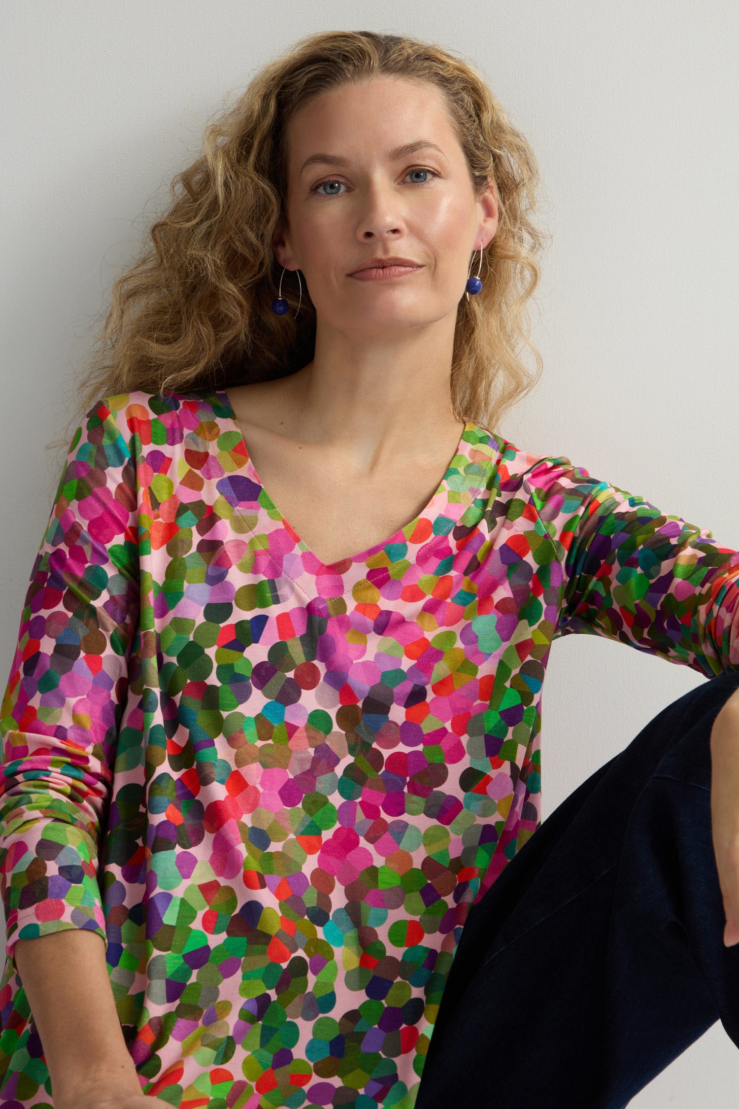 Person with long curly hair wears a V-neck Abstract Confetti Jersey Top while seated against a plain background, looking at the camera.