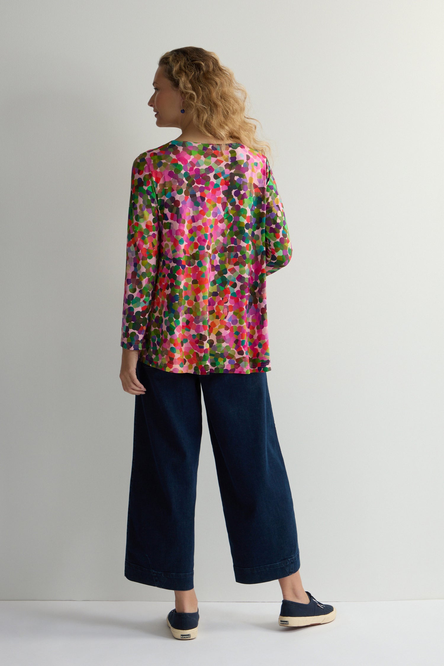 A person stands with their back to the camera, wearing a V-neck Abstract Confetti Jersey Top, paired with wide-leg denim pants and dark slip-on shoes against a simple backdrop.