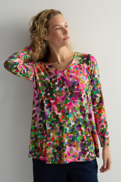 Wearing an Abstract Confetti Jersey Top with a V-neck and fit-and-flare design, the person stands against a plain background, hand touching hair.