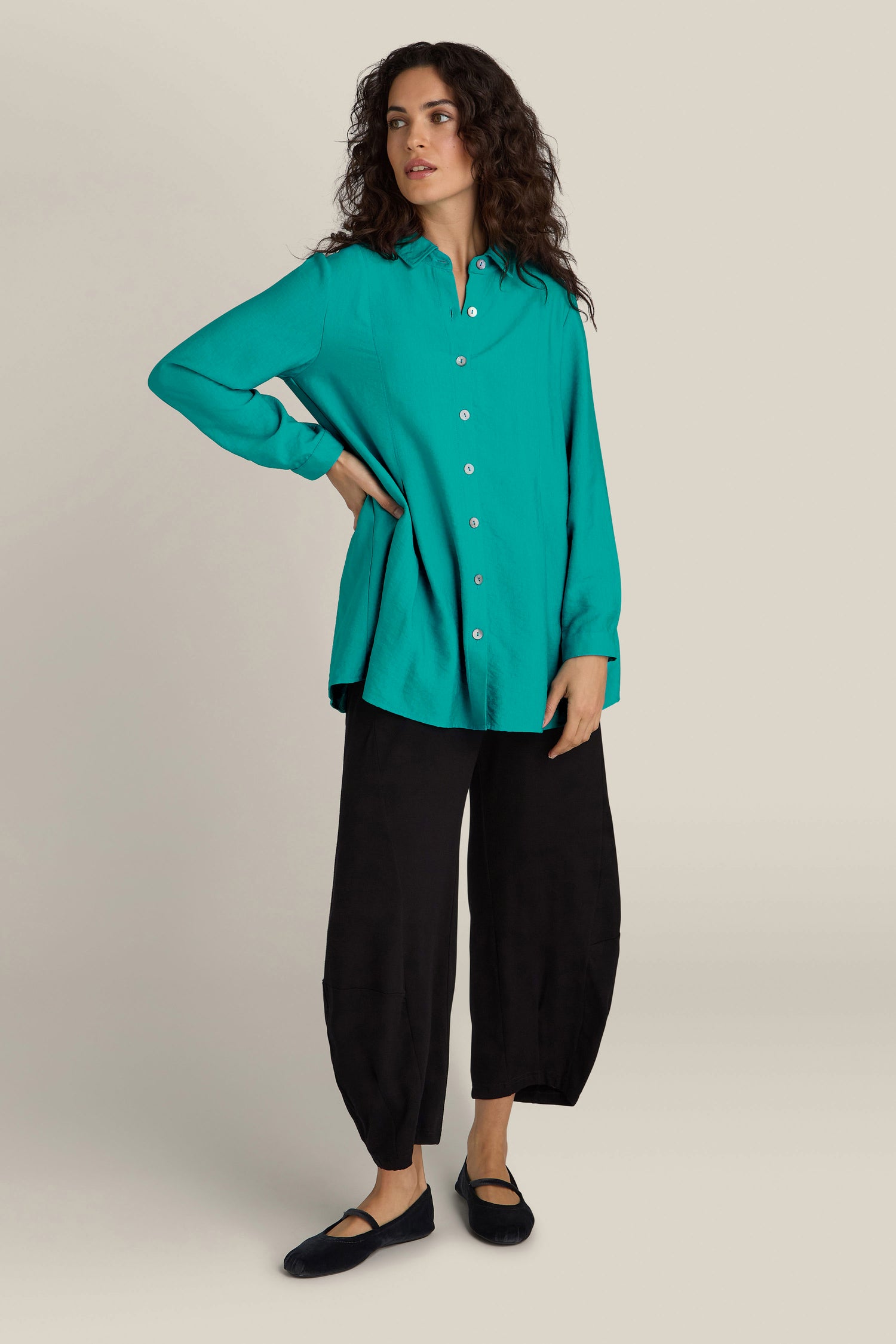 A person in a Textured Viscose Pleat Shirt, turquoise blue in color, and black loose pants stands against a plain background. They have relaxed, curly hair and wear black shoes. The silky textured viscose fabric of the shirt adds an elegant contemporary touch to their look.