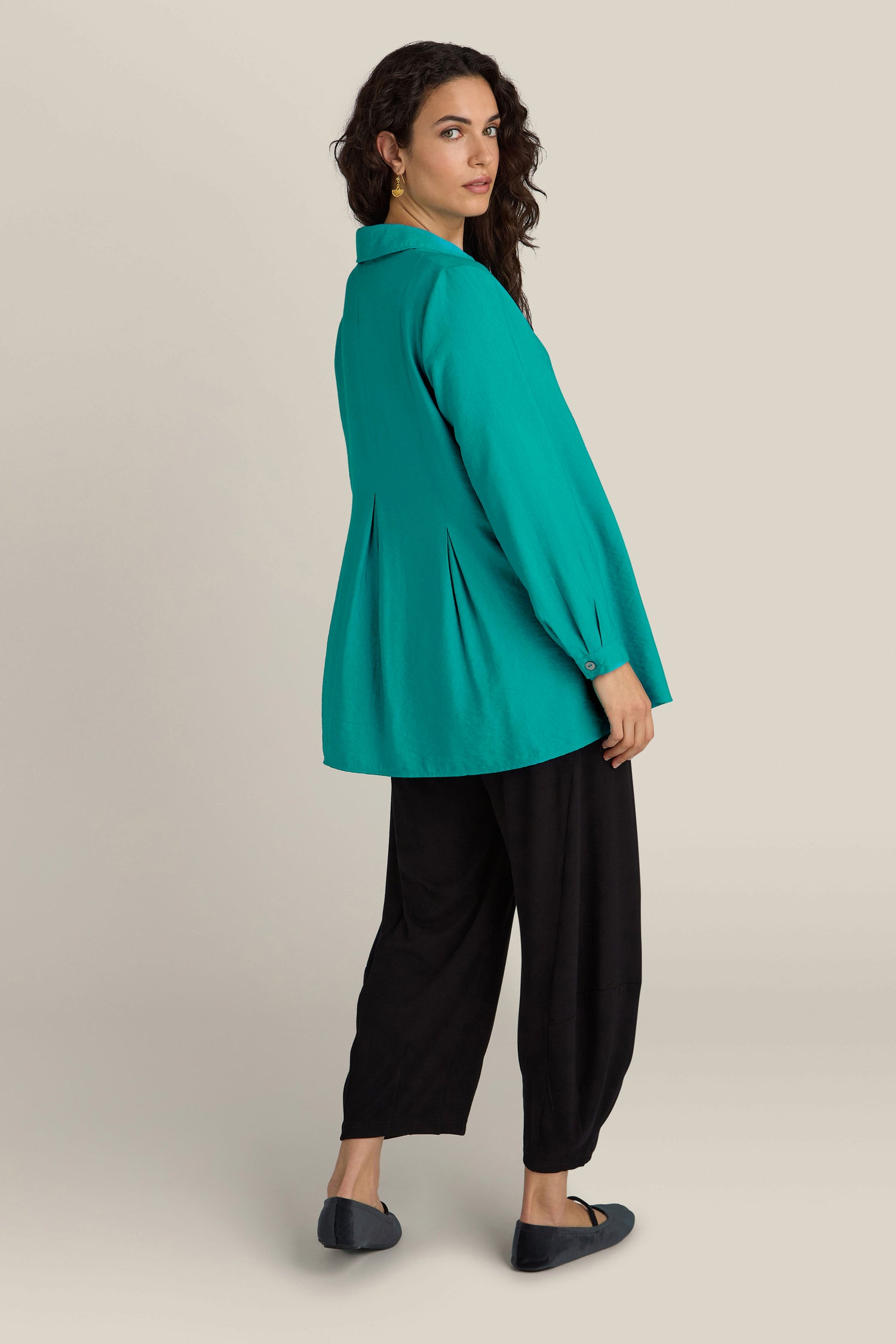 Person wearing a loose, long-sleeved Textured Viscose Pleat Shirt in teal over black pants and black flat shoes, standing on a light gray background and looking over their shoulder to the left. The silky textured viscose fabric of the shirt adds an elegant touch to the contemporary ensemble.