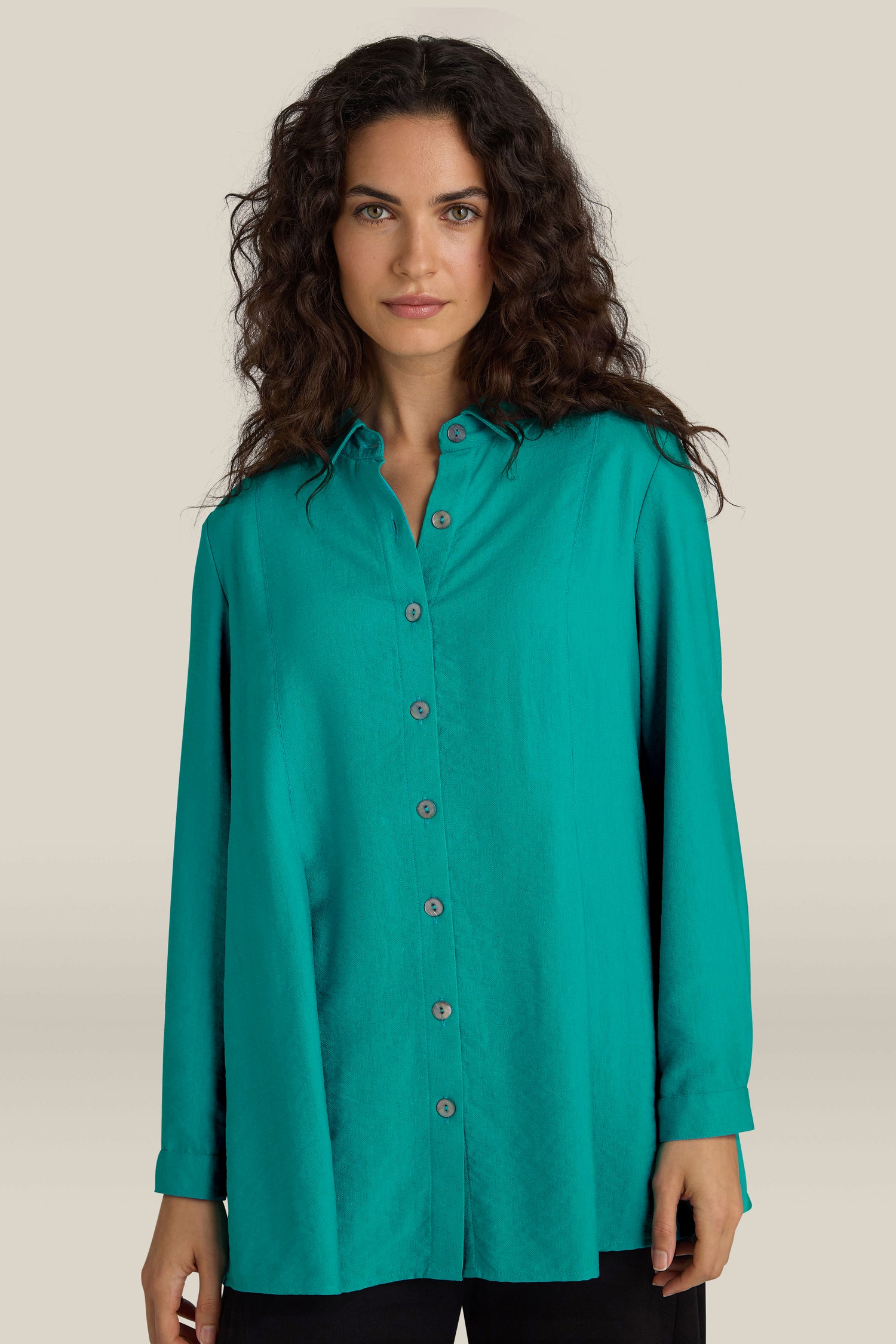 A woman with long, curly hair is wearing an elegant Textured Viscose Pleat Shirt in turquoise blue and black pants, standing in front of a plain beige background.