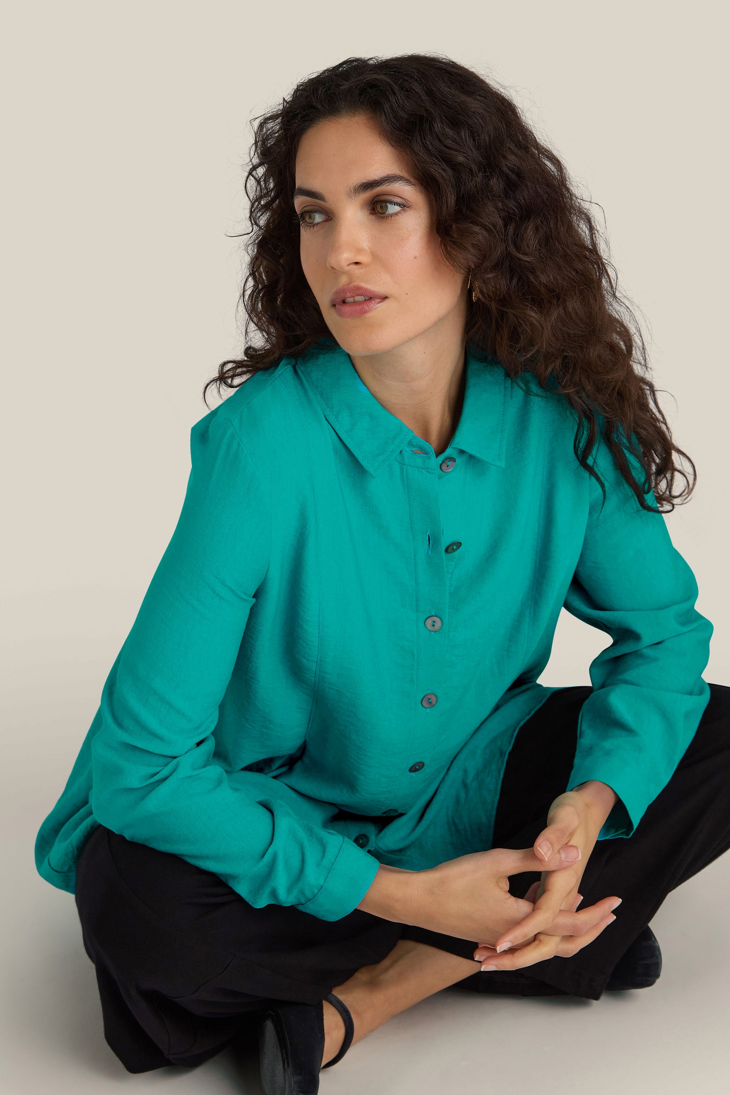 A woman with curly hair, wearing a turquoise blue Textured Viscose Pleat Shirt and black pants, sits on the floor with crossed legs and looks to the side.