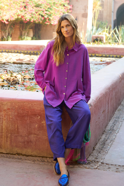 A person sits on the edge of a pink stone fountain, wearing the Textured Viscose Pleat Shirt in bright purple and blue pants, with plants and flowers in the background.