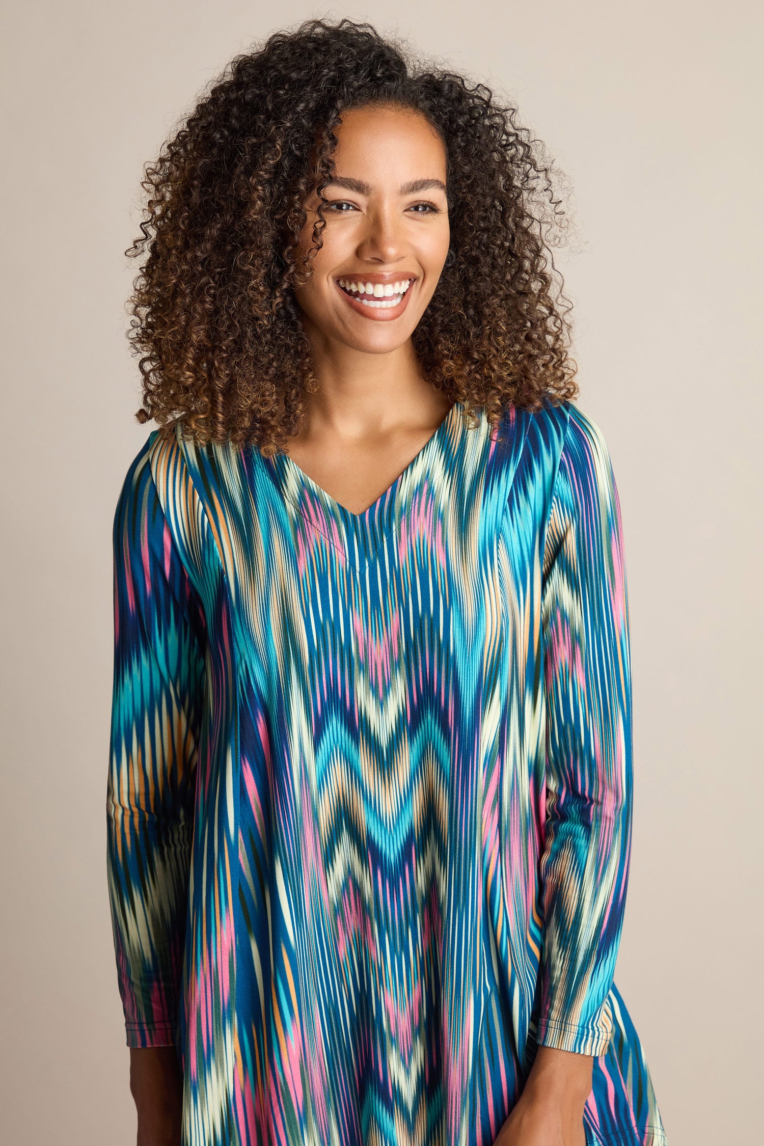 A woman with curly hair, wearing the vibrant Autumnal Ikat Jersey Top featuring a colorful pattern, smiles while standing against a plain background.