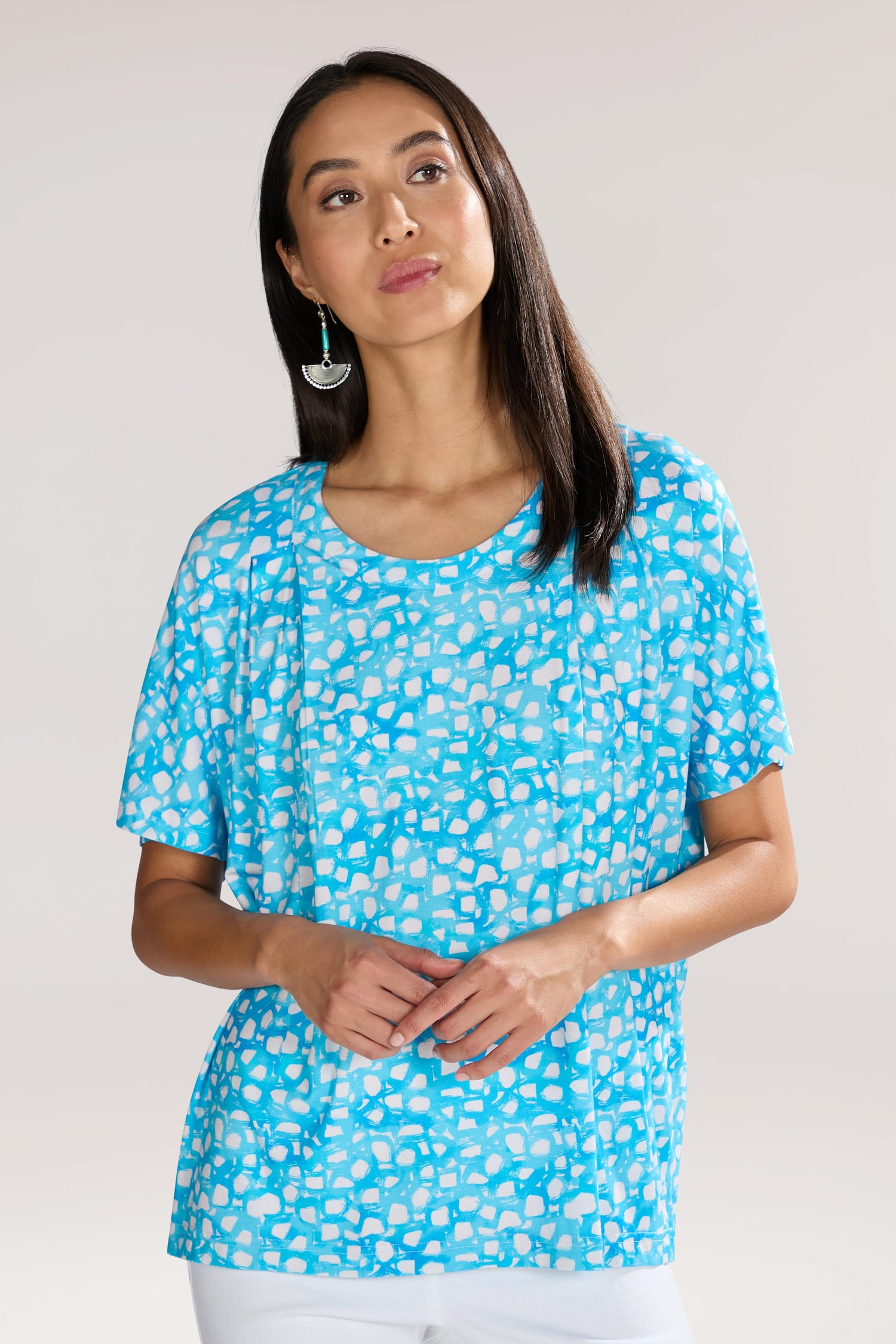 A woman with long dark hair wearing a blue and white Pebble Jersey Print Relaxed Top with an abstract bubble motif stands against a plain background, her earrings adding a touch of elegance.