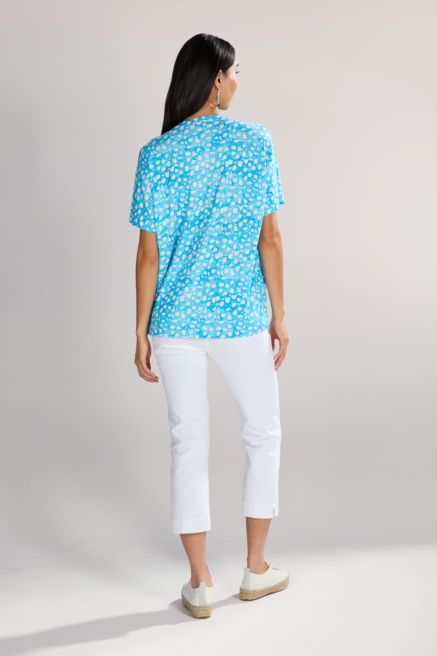 A person with long dark hair is standing with their back to the camera, wearing a Pebble Jersey Print Relaxed Top featuring an abstract bubble motif, white pants, and white shoes against a neutral background.