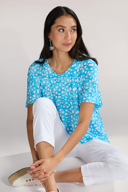 A woman with long dark hair sits on the floor, wearing a blue Pebble Jersey Print Relaxed Top with an abstract bubble motif, white pants, and beige sneakers. She is looking to the side with a neutral expression.