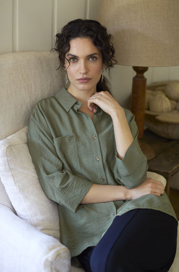 A person with curly hair sits on a beige couch wearing a relaxed chic, green Textured Viscose Boxy Shirt and dark pants, with one hand touching their face.