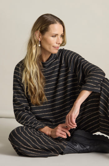 A person with long, wavy hair sits on the floor, wearing a black Broken Stripe Jersey Top detailed with gold horizontal stripes and a mock neck, gazing to the side.