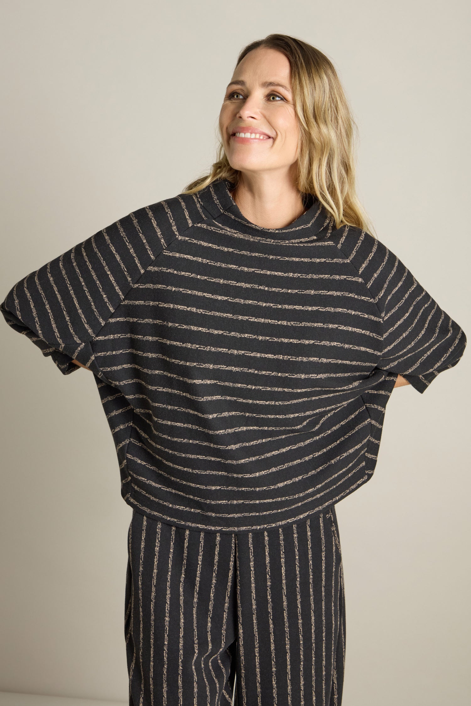 A woman dressed in a Broken Stripe Jersey Top with dolman sleeves and coordinating pants stands against a plain background, gazing upward and smiling.