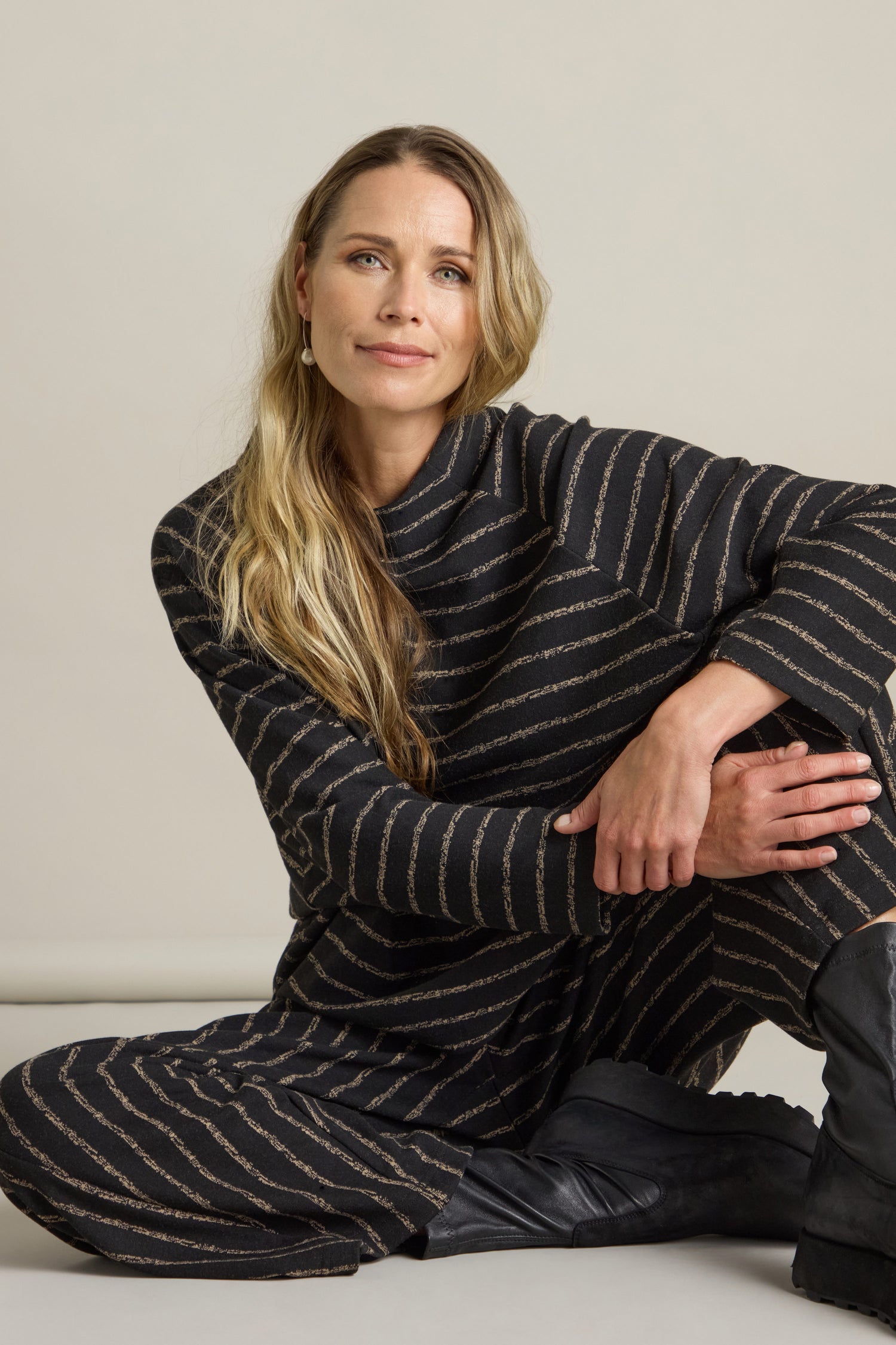 A woman with long blonde hair, wearing the Broken Stripe Jersey Top featuring dolman sleeves and a mock neck, sits on the floor against a plain background. She rests one arm on her knee and looks directly at the camera.