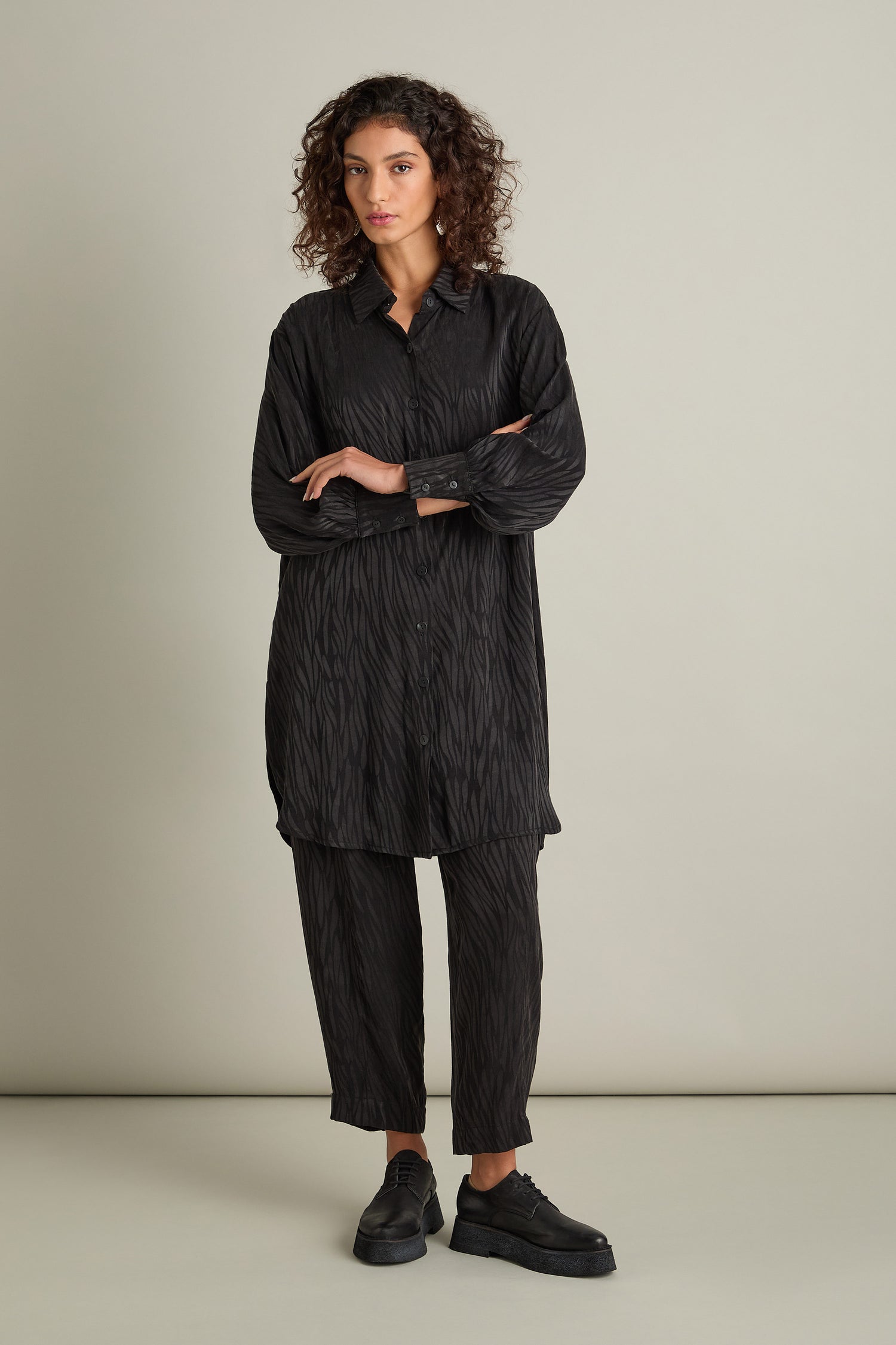 A person with curly hair stands against a plain background, wearing an Abstract Lines Jacquard Long Shirt in black, showcasing its relaxed fit and silky-feel fabric texture, paired with matching black pants and black shoes.