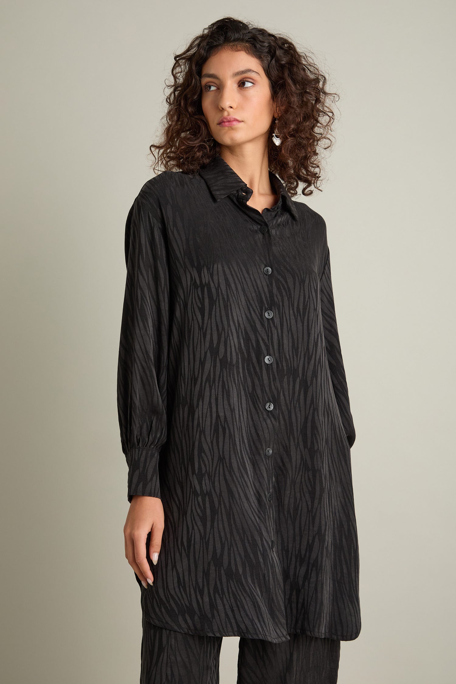 A person with curly hair is wearing an Abstract Lines Jacquard Long Shirt, featuring long sleeves and a button-up design in a silky-feel fabric, standing against a plain background.