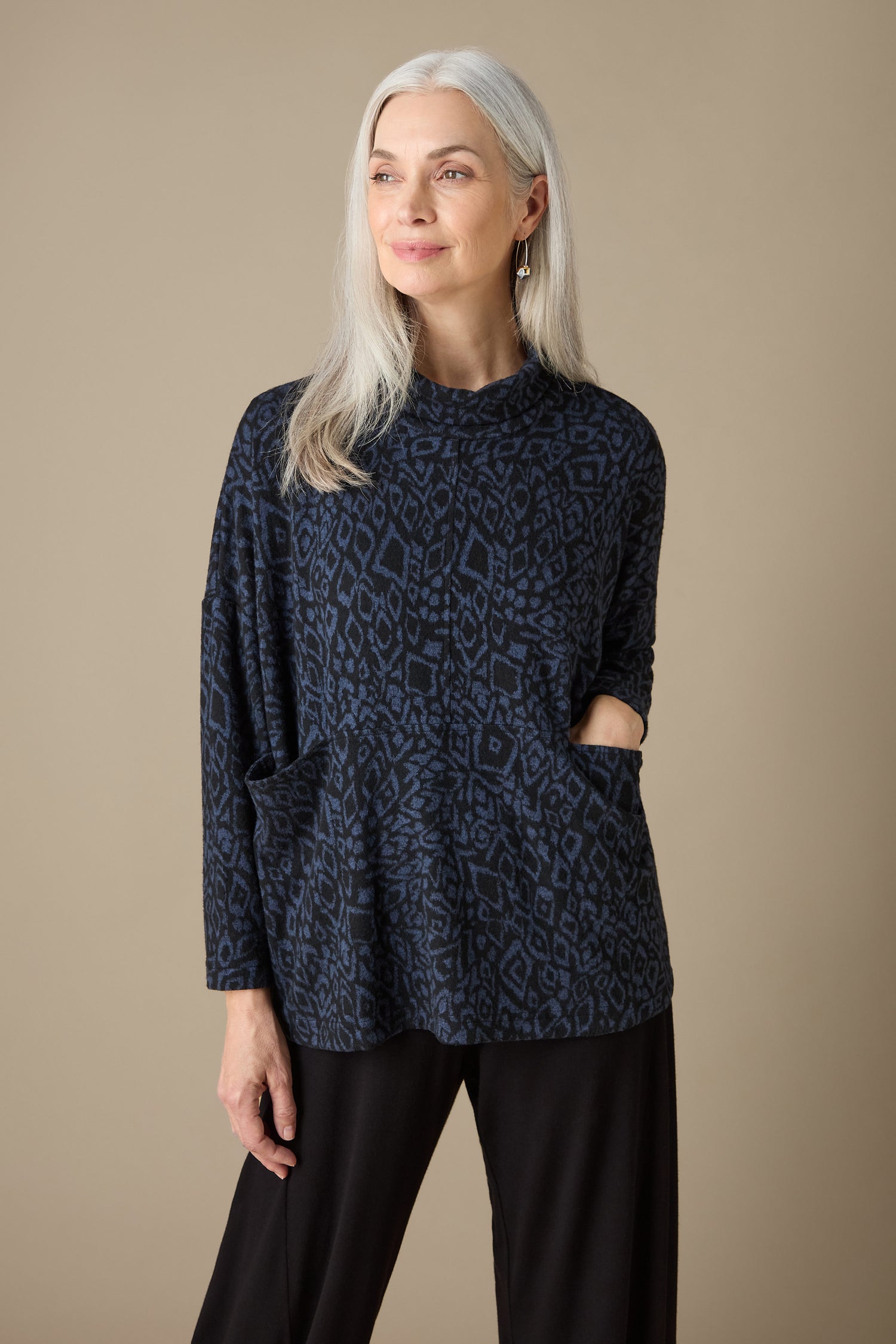 A woman with long gray hair is wearing the Ikat Soft Jersey Relaxed Top, which features an abstract print in dark tones, paired with black pants. She stands gracefully against a plain beige background.