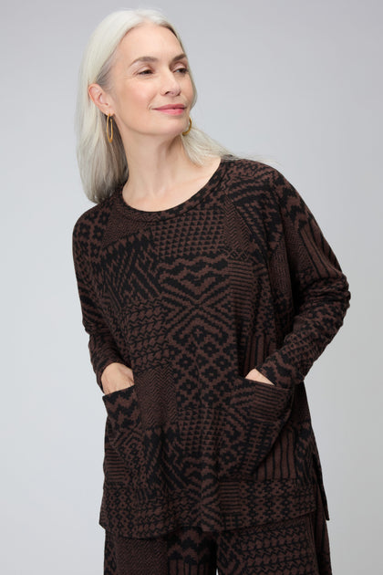 A woman with long gray hair, gold hoop earrings, and an oversized Textured Ikat Patchwork Jersey Top with pockets stands against a plain light background.