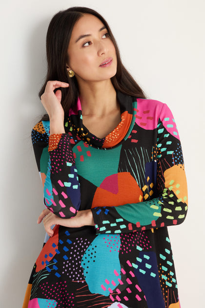 A woman with long dark hair models the Mark Making Print Jersey Tunic, featuring a colorful artistic pattern, as she stands against a white wall and looks upward with her hand touching her hair.