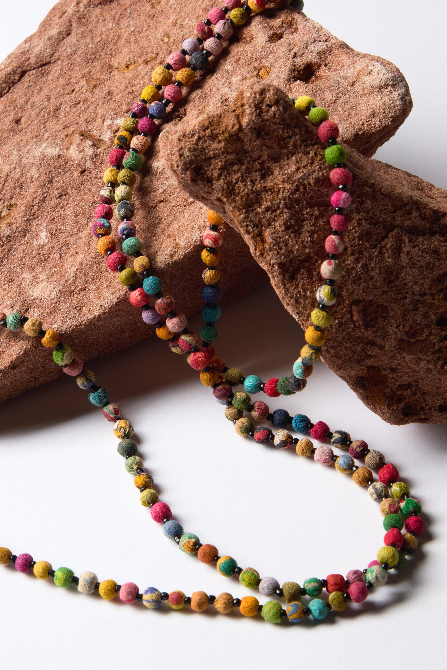 The Kantha Long Bead Spheres Necklace showcases colorful strands of fabric beads, elegantly arranged over two large brown stones on a white surface. Crafted from repurposed textiles, it offers a vibrant blend of colors and textures.