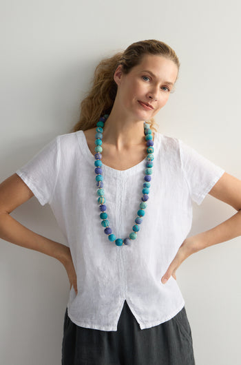 Against a plain background, a person with hands on hips wears a white blouse and the vibrant Kantha Garland Necklace, handcrafted from repurposed textiles, adding an artistic flair to their simple attire.