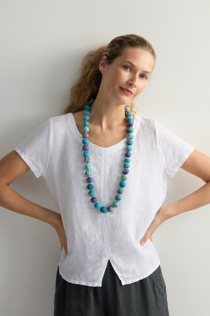 Against a plain background, a person with hands on hips wears a white blouse and the vibrant Kantha Garland Necklace, handcrafted from repurposed textiles, adding an artistic flair to their simple attire.