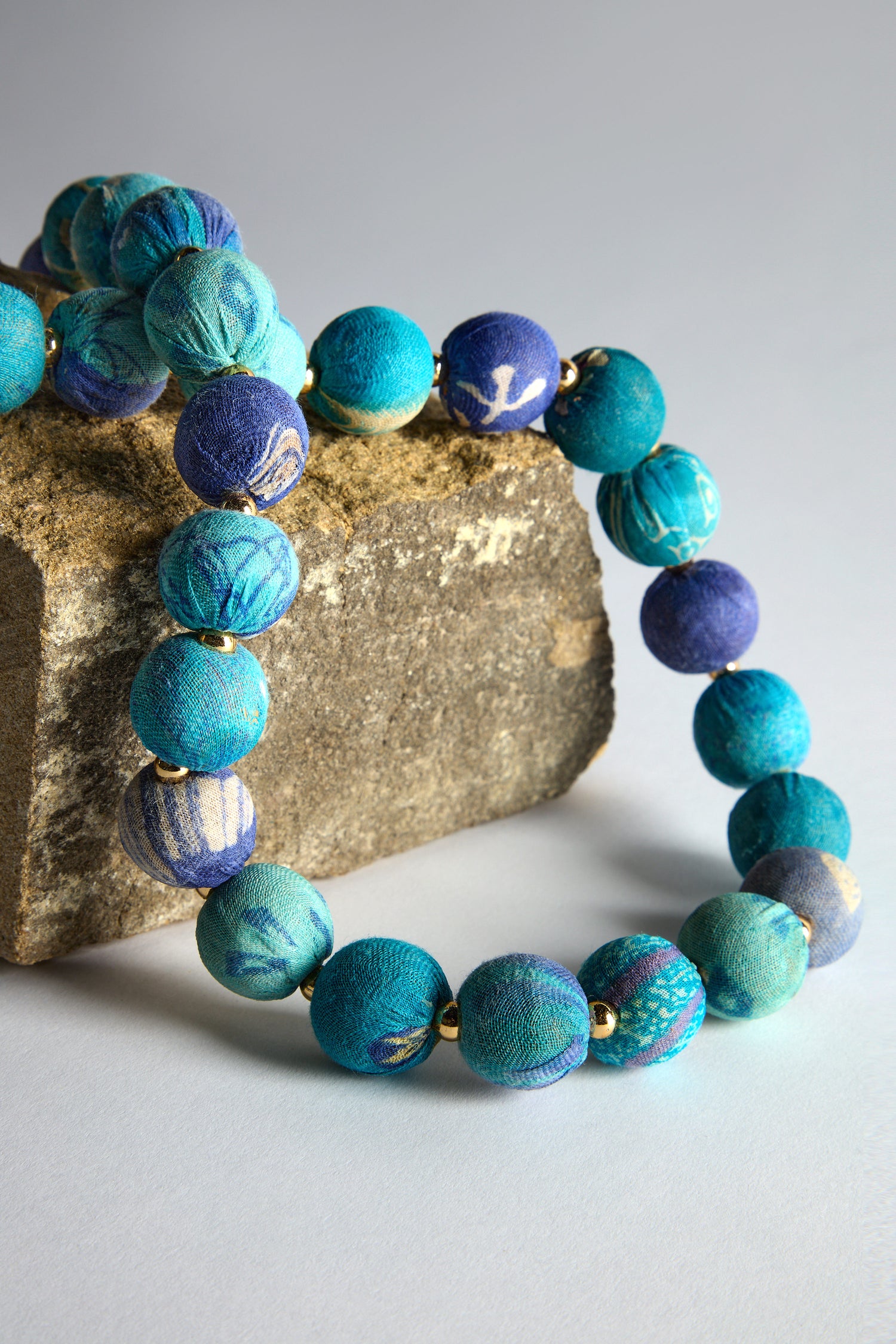 The Kantha Garland Necklace, featuring handcrafted blue and teal fabric-covered beads with subtle patterns made from repurposed textiles, is elegantly draped over a rough stone block.