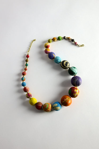 The Kantha Graduated Bead Necklace, made from recycled sari fabric, features large, colorful beads in blue, red, and yellow arranged in an S-shape on a light background.