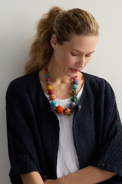 A woman with a ponytail, wearing a textured black jacket, white shirt, and a Kantha Graduated Bead Necklace made from recycled sari fabric, looks down against a plain backdrop.