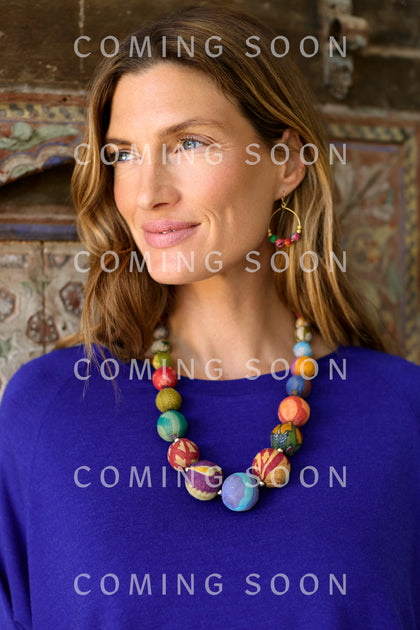 A woman with wavy hair in a blue top and geometric earrings wears the Kantha Graduated Bead Necklace, standing before a decorative background. "COMING SOON" and "KANTAC751" appear multiple times over the image.