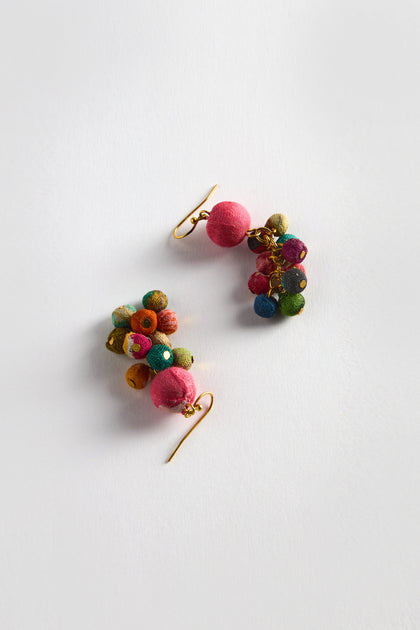 The Kantha Spheres Earrings are vibrant, featuring clusters of multi-colored textile spheres with gold hooks. They are meticulously handcrafted from recycled textiles, set against a white background.