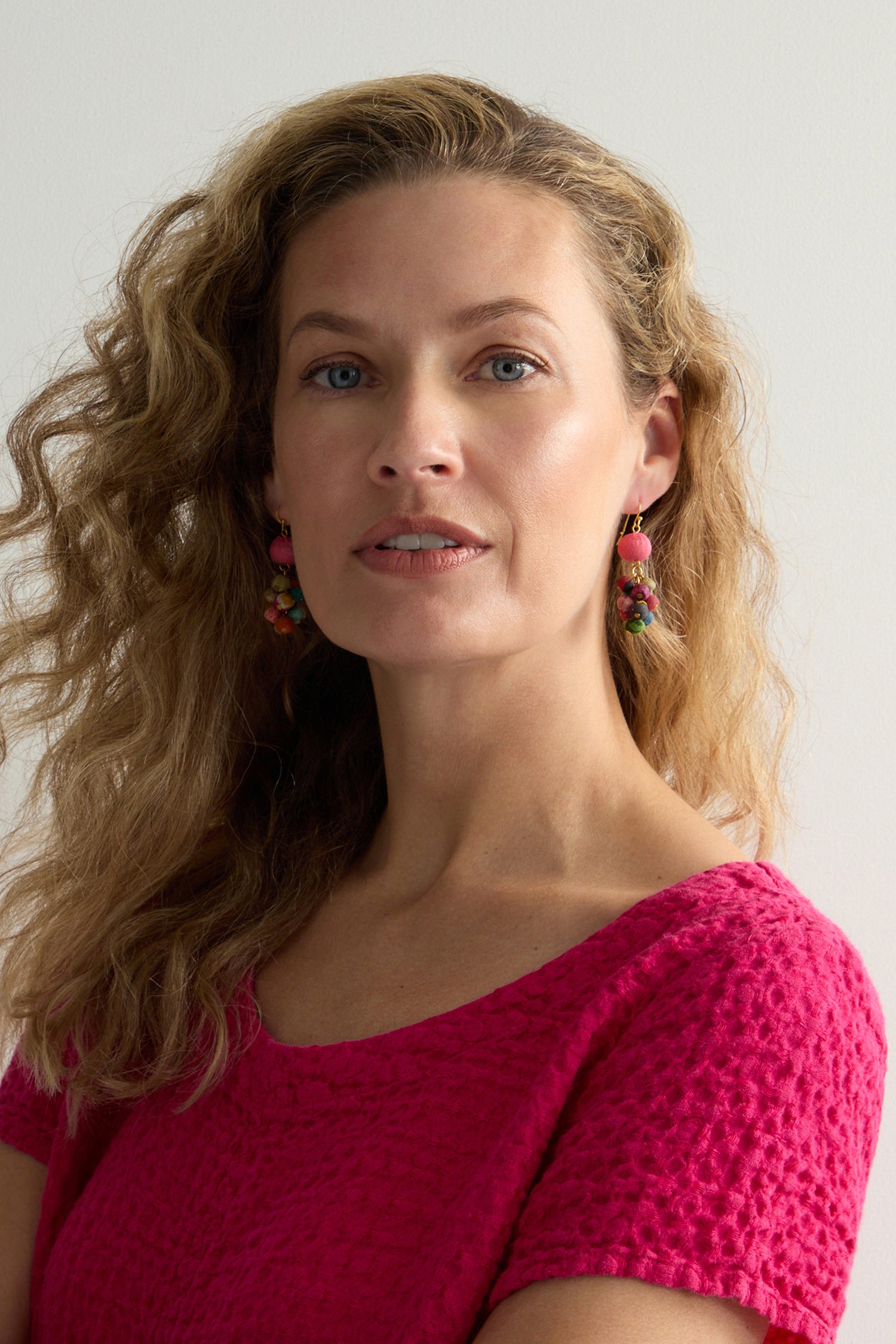 A woman with wavy blonde hair in a pink textured top stands against a neutral background, showcasing the colorful Kantha Spheres Earrings made from recycled textiles.