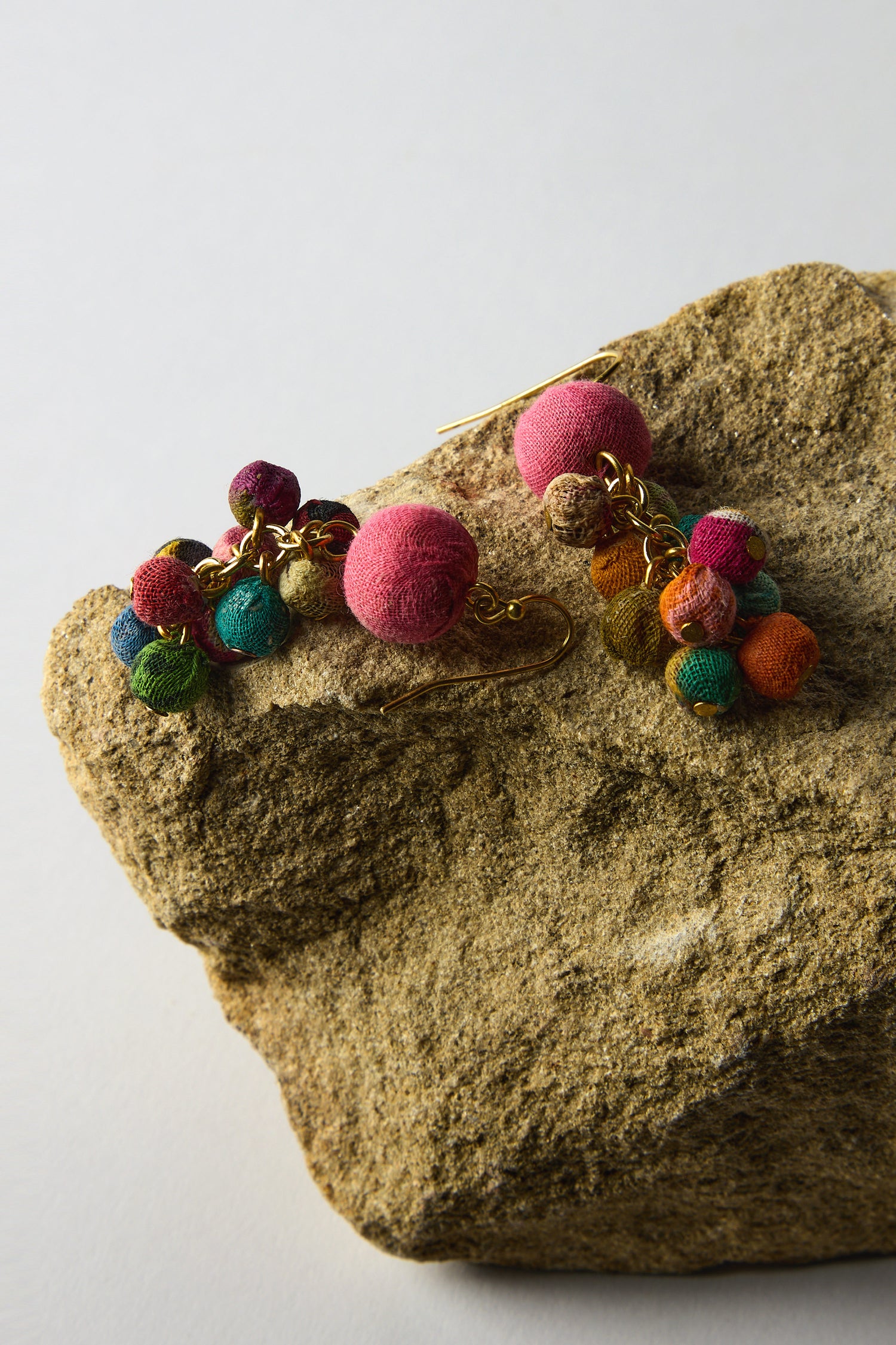 Kantha Spheres Earrings, made from recycled textiles, feature vibrant beads with gold hooks elegantly placed on a textured rock surface.