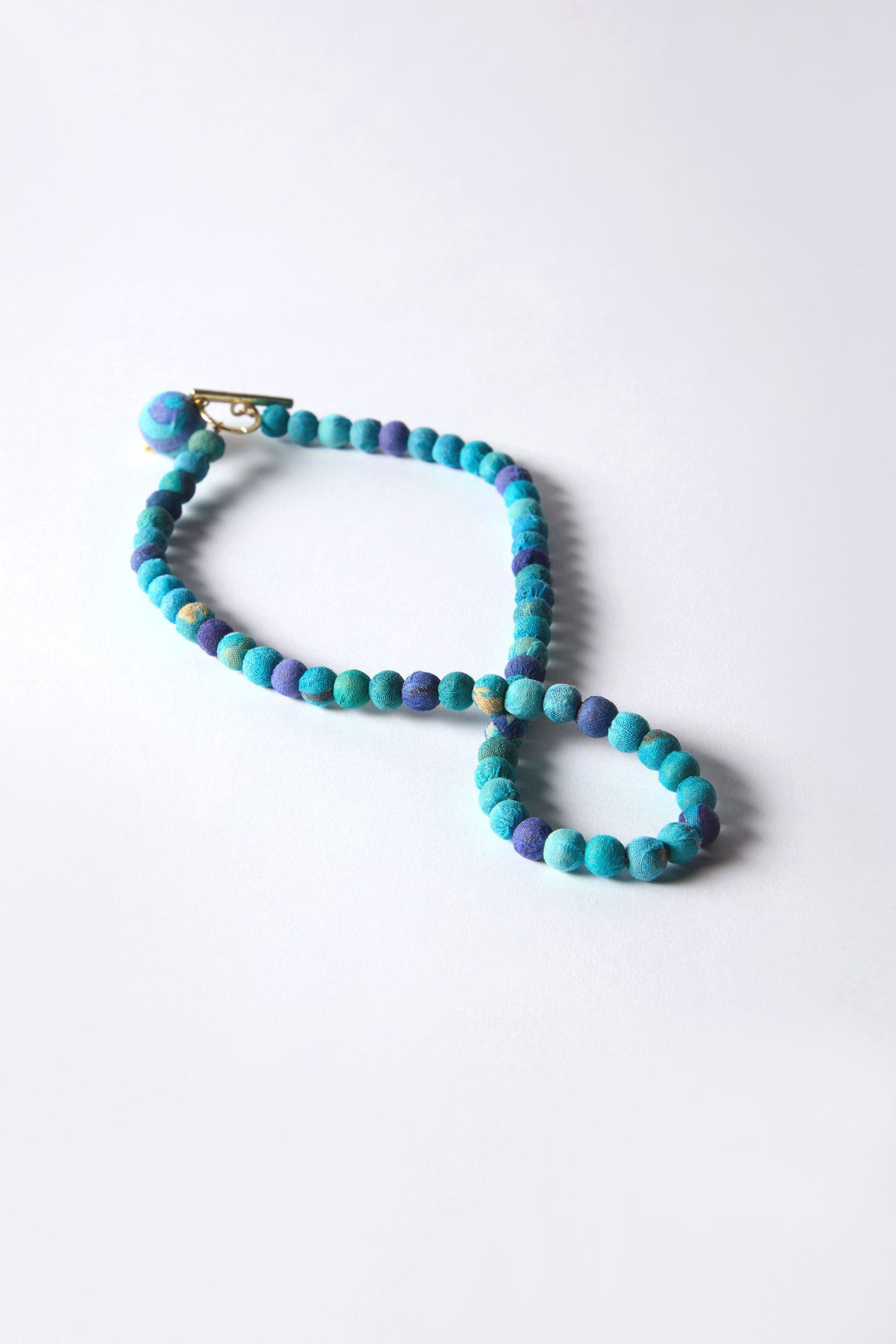 Displayed on a white surface, the Kantha Toggle Necklace in blue and purple beads highlights artisanal craftsmanship with repurposed textiles and a simple clasp.