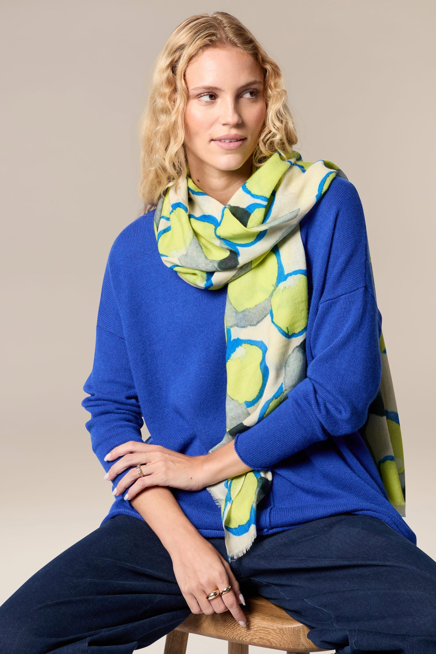 A woman is sitting on a stool wearing a blue sweater and the Neon Spot Wool Scarf, which serves as a statement accessory for her all-occasion styling.