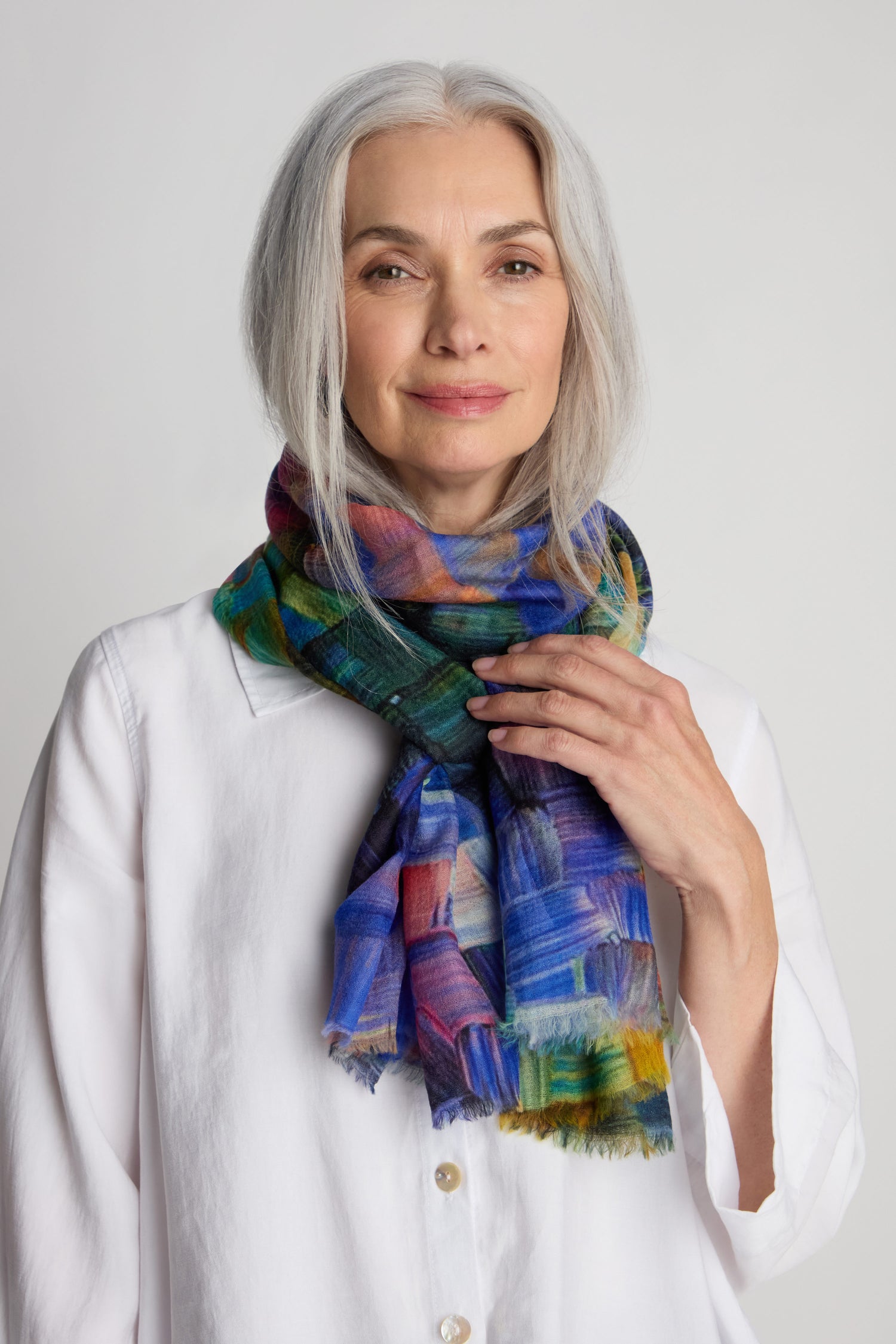 A woman with gray hair wears a white button-up shirt and a Painterly Merino Wool Scarf that resembles hand-painted art. She is looking at the camera with a neutral expression.