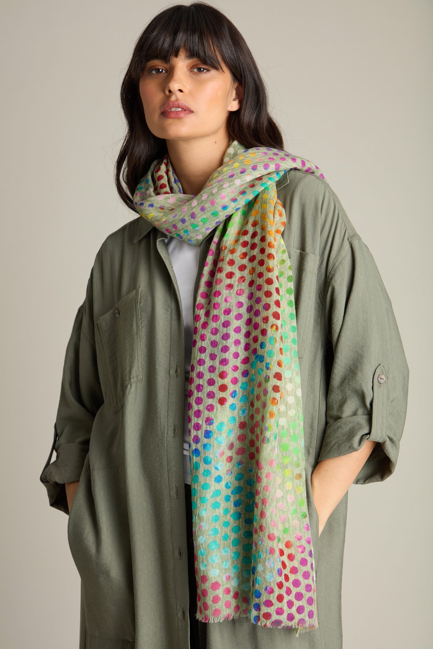 A person wearing a green jacket and a vibrant accessory, the Mini Rainbow Spot Merino Wool Scarf, standing against a plain background.