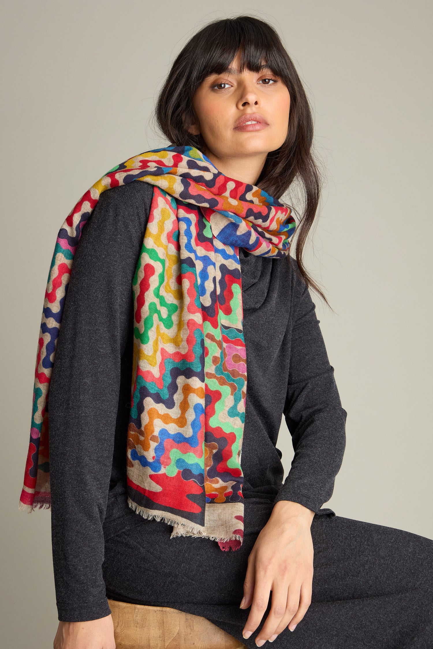 A person wearing a dark outfit and a colorful, patterned Reverie Print Wool Scarf, crafted from premium soft wool, sits on a wooden stool against a plain background.