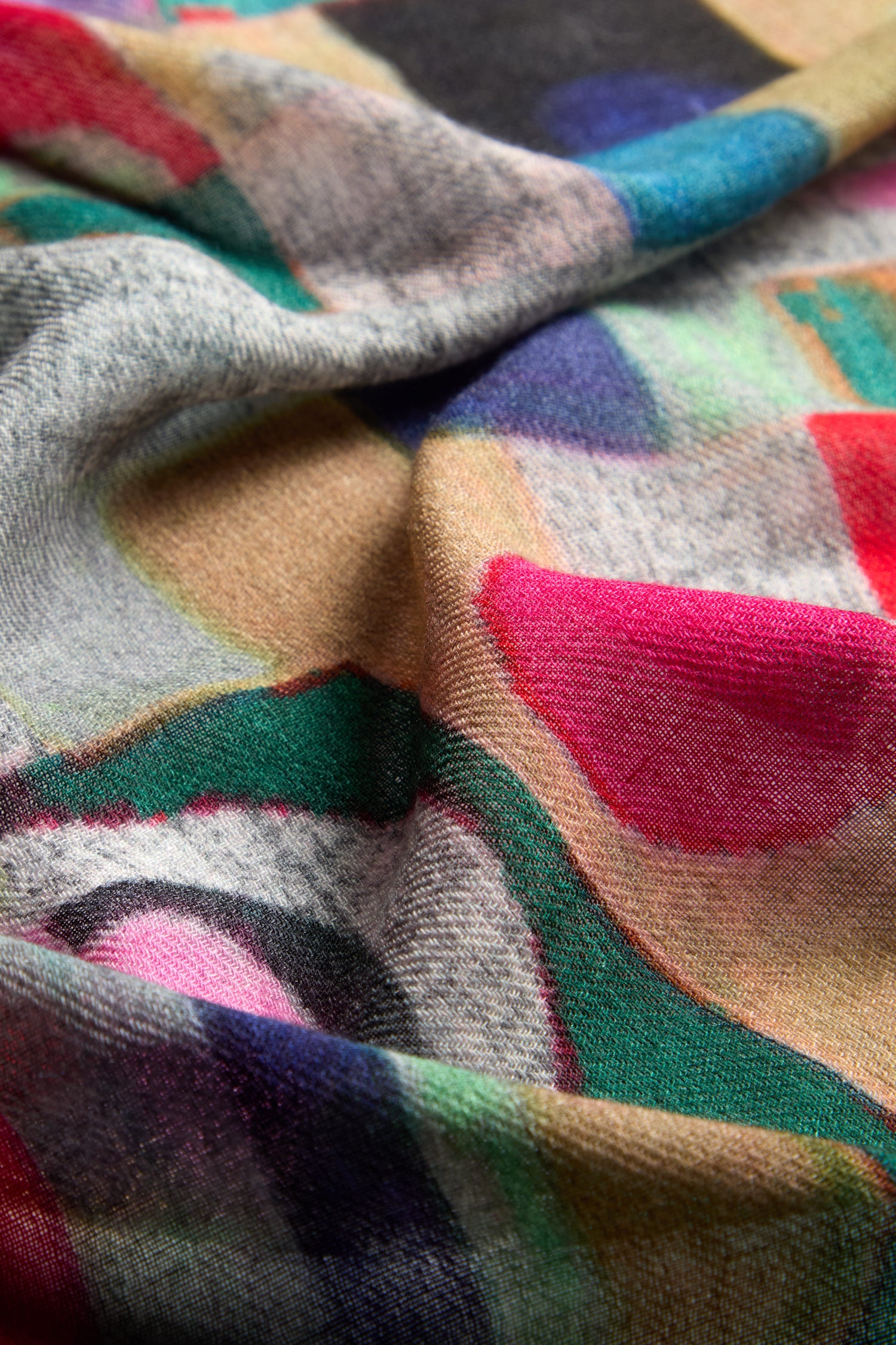 A close-up view of the Kandinsky Merino Wool Scarf, which features a multicolored, abstract-patterned fabric reminiscent of Kandinsky's abstract expressionism. It showcases vivid patches of pink, green, blue, and beige and appears soft with a slightly textured surface.